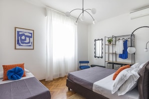 A bedroom with two beds, a hanging light fixture, a mirror, a clothing rack, and a framed artwork on the wall.