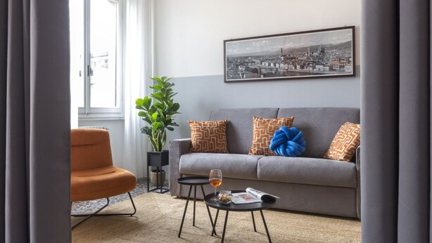 Una sala de estar con un sofá gris, un sillón naranja, una mesa de centro negra con una copa de vino, una planta en maceta y un cuadro con una vista urbana en la pared.