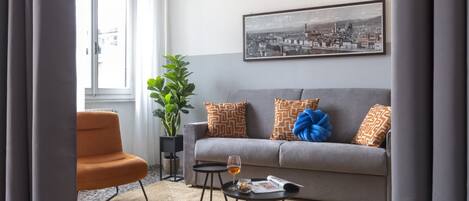 A living room with a grey sofa, orange armchair, a black coffee table with a glass of wine, a potted plant, and a framed cityscape picture on the wall.