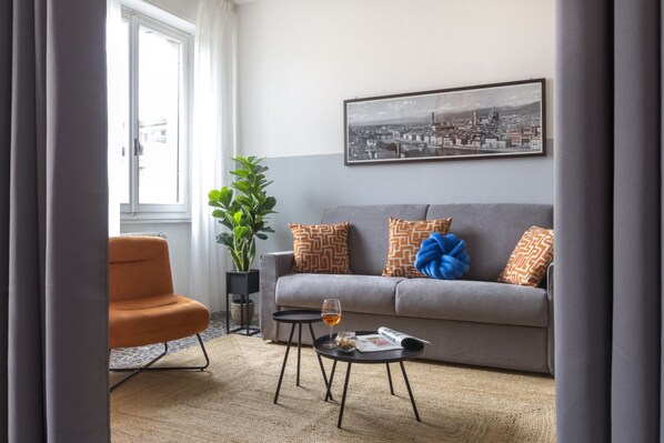 Un salon avec un canapé gris, un fauteuil orange, une table basse noire sur laquelle se trouve un verre de vin, une plante en pot et, au mur, un tableau encadré représentant un paysage urbain.