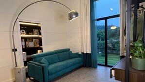 A teal sofa in a modern living room with a built-in bookshelf, a large window with a view of greenery, and a compact, dark-colored side table. - Mille912 relais: Deluxe apartment  (Piano di Sorrento)