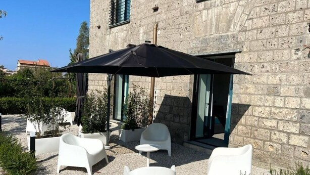 A courtyard with white plastic chairs and a large black umbrella, set against a stone building with a window and a door. - Mille912 relais: Deluxe apartment  (Piano di Sorrento)