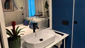 A modern bathroom with a white sink, a large mirror, and a view of a bedroom with a bed and a window. - Mille912 relais: Deluxe apartment  (Piano di Sorrento)