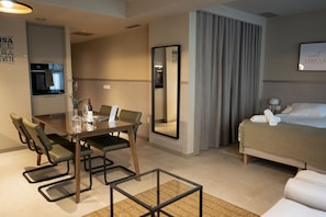 A hotel room with a bed, a dining table with chairs, a kitchen area, and a large mirror.
