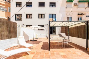 A courtyard with white lounge chairs, a covered seating area, and a building with multiple windows in the background.