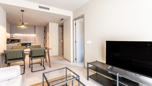A modern living room with a flat-screen TV, a glass coffee table, and a sofa. There is a kitchen area with a dining table and chairs.