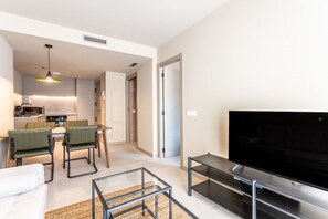 A modern living room with a flat-screen TV, a glass coffee table, and a sofa. There is a kitchen area with a dining table and chairs.