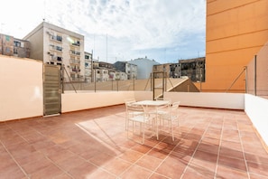 A rooftop terrace with a table and chairs, a metal fence, and buildings in the background.