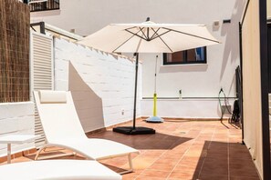 Une terrasse ensoleillée avec un fauteuil de jardin blanc, un grand parasol et une fenêtre donnant sur un beau paysage.