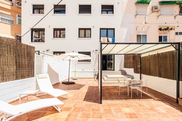 A courtyard with white lounge chairs, a covered seating area, and a building with multiple windows in the background.