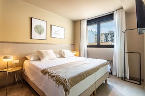 A hotel room with a large bed, two bedside tables with lamps, a window with curtains, and two framed pictures on the wall.