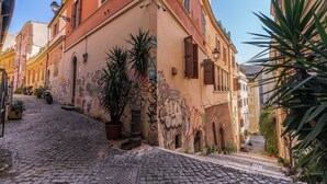 A cobblestone street with buildings featuring arched windows and balconies, some with shutters, and graffiti on the walls. - CIANCALEONI APPARTAMENTO (Rome)
