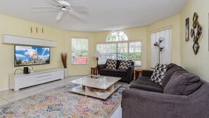 A living room with a flat-screen TV mounted on the wall, a ceiling fan, a sofa, a coffee table, and a patterned rug. - Villas at Island Club Community, Pools, Jacuzzi and More! (Kissimmee)