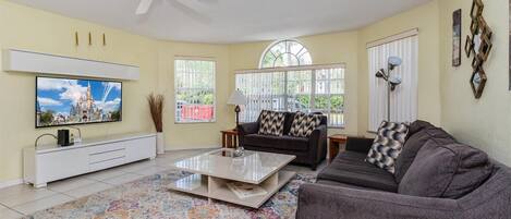 A living room with a flat-screen TV mounted on the wall, a ceiling fan, a sofa, a coffee table, and a patterned rug.