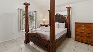 A four-poster bed with a wooden headboard, a nightstand with a lamp, and a wooden dresser with drawers. - Cozy Home Near Disney!!! (Kissimmee)