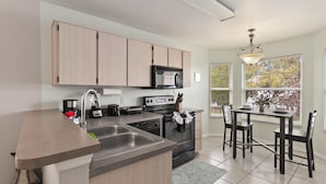 A modern kitchen with light wood cabinets, a stainless steel sink, a microwave, an oven, and a dining area with a table and chairs. - Cozy Home Near Disney!!! (Kissimmee)
