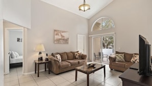 A spacious living room with a sofa, coffee table, and a TV. There is a door leading to another room and a window with a view of the outdoors. - Cozy Home Near Disney!!! (Kissimmee)