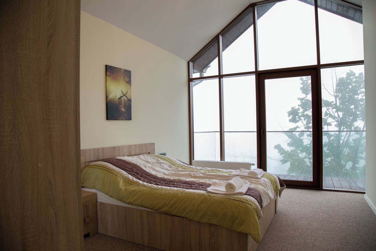 Un dormitorio con un ventanal grande, una cama con colcha verde y beige, cabecero de madera y un cuadro en la pared.