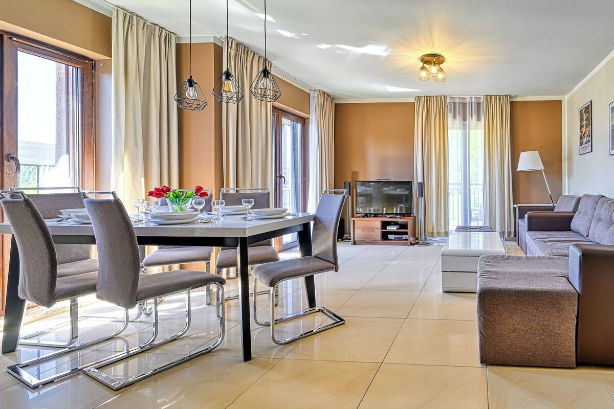 Una habitación de hotel moderna con zona de comedor, sofá, televisor y un cuadro en la pared.