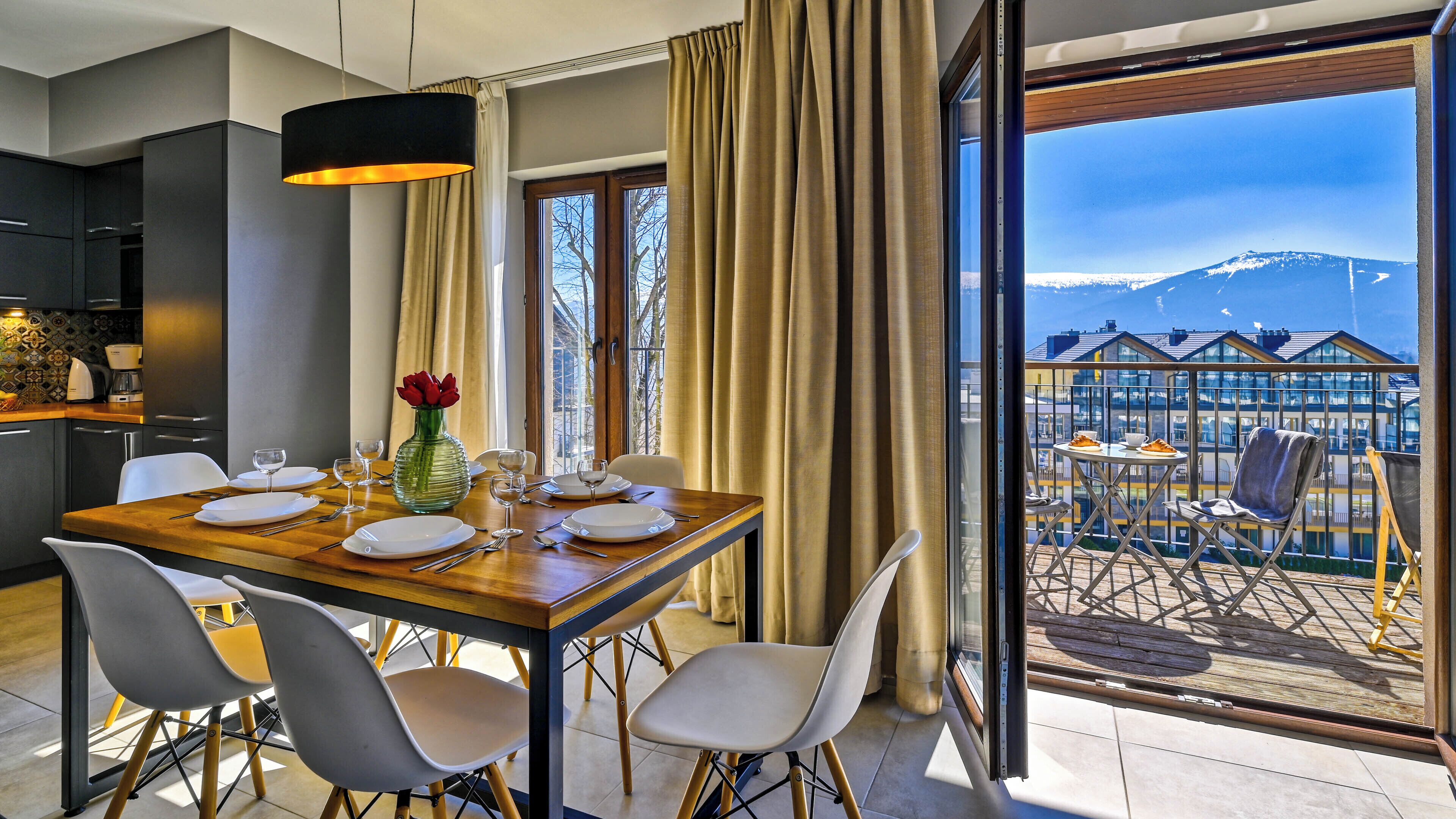 A dining area with a table set for four, a vase with red flowers, and a view of a mountain through large windows.