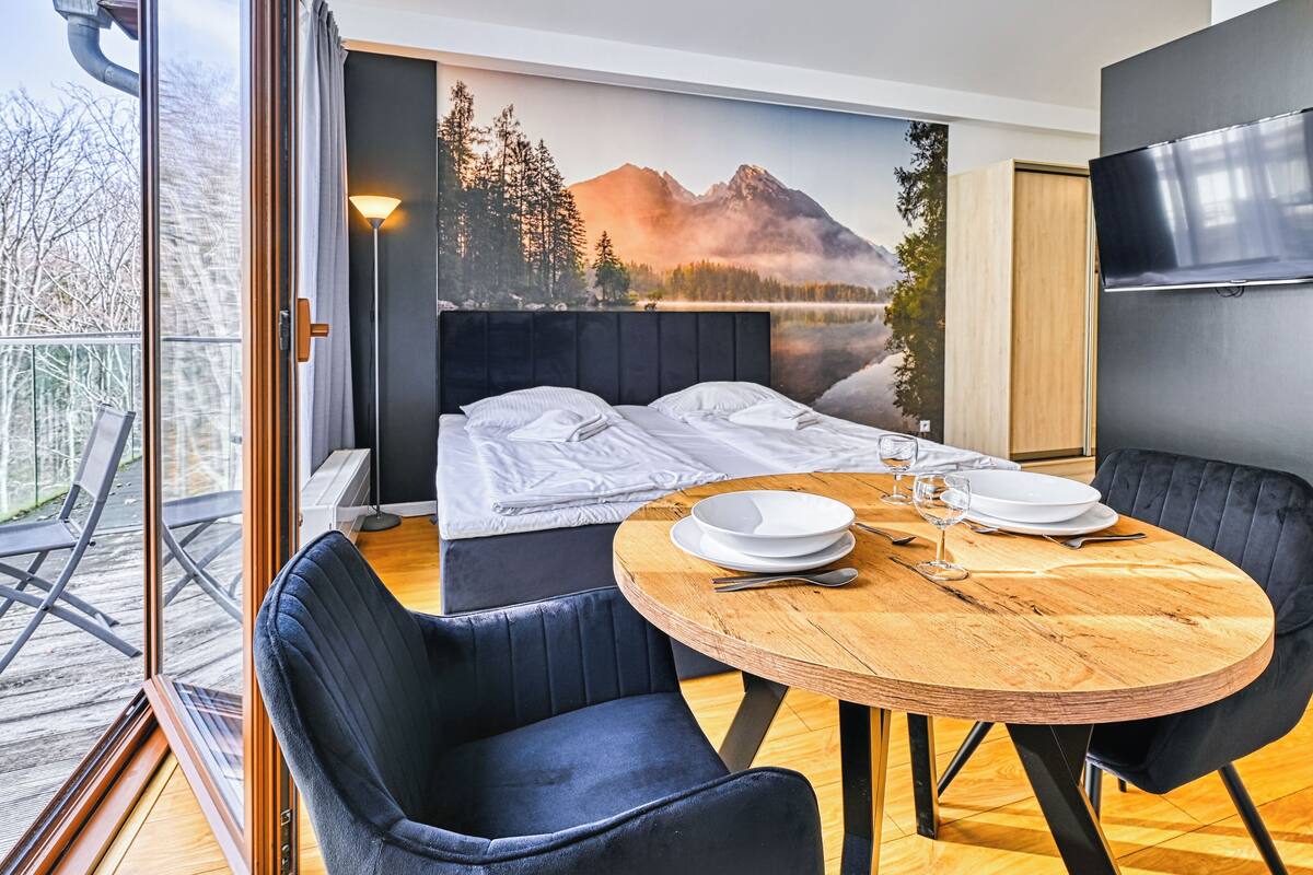 Habitación de hotel moderna con cama, una mesa de comedor dispuesta para dos personas y un amplio ventanal con vistas a un paisaje montañoso.