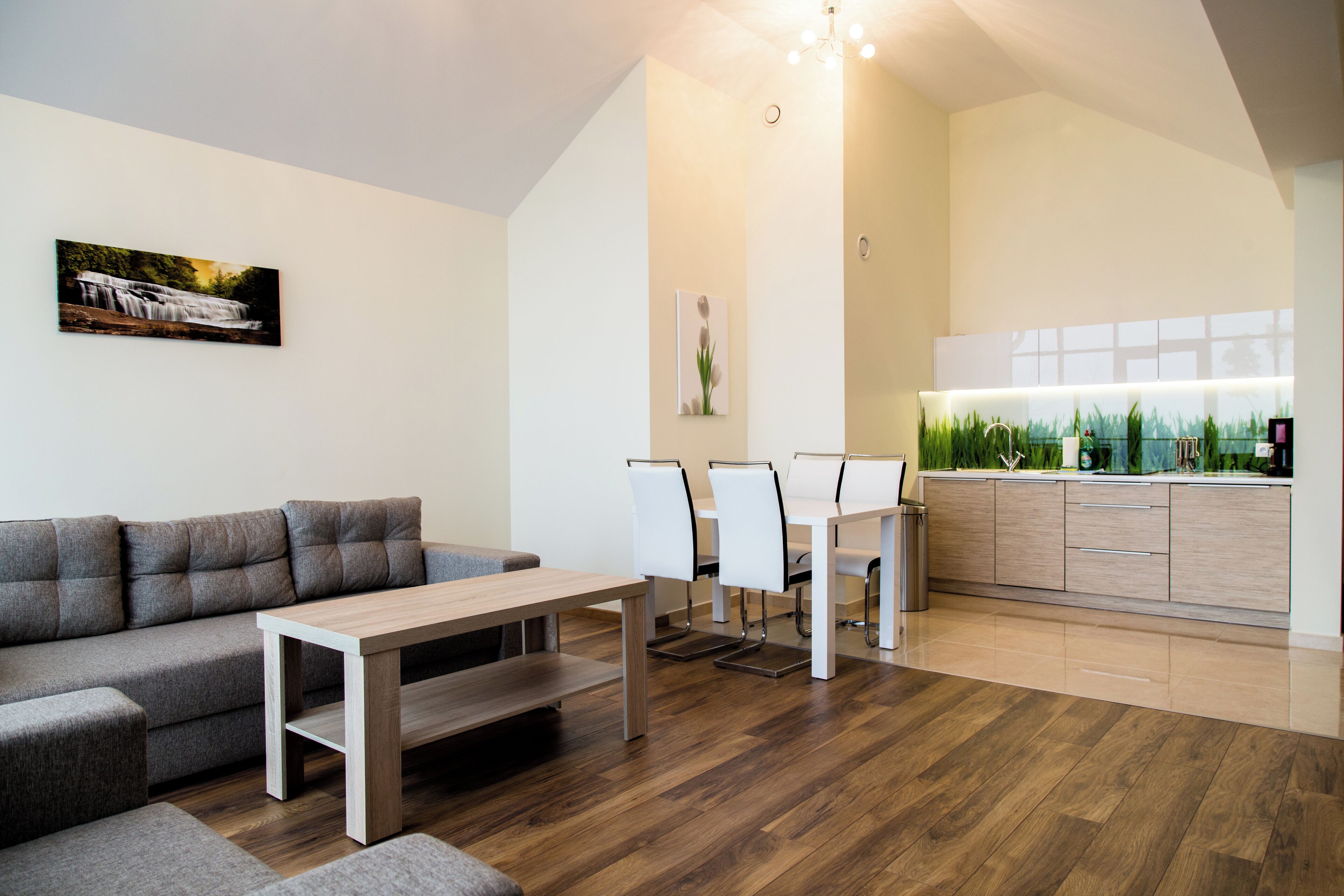 A modern living room with a grey sofa, a wooden coffee table, and a dining area with white chairs and a glass-top table.