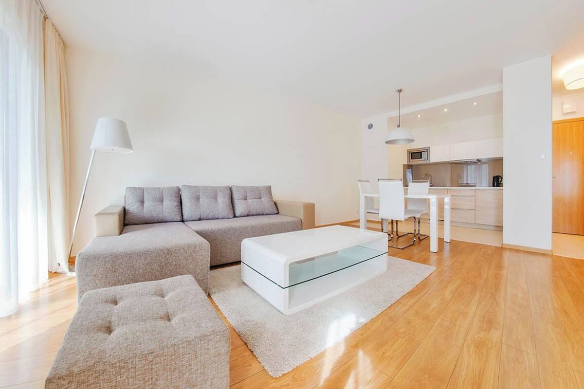 Una sala de estar moderna con sofá, reposapiés y mesa de centro de vidrio. Se observa una cocina al fondo.