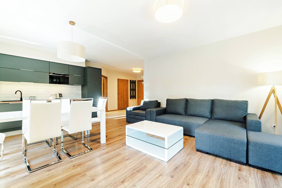 Una sala de estar moderna con un sofá gris, una mesa de centro blanca y una zona de comedor con sillas blancas, y una cocina al fondo.
