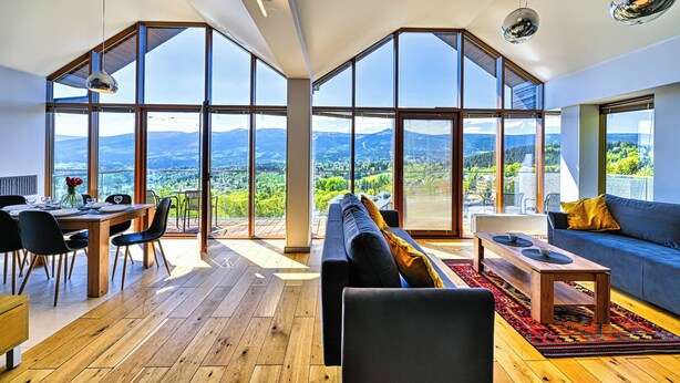 Amplia sala de estar con ventanas grandes que ofrecen vistas a la montaña, una mesa de comedor de madera y una zona de estar acogedora con un sofá y una mesa de centro.
