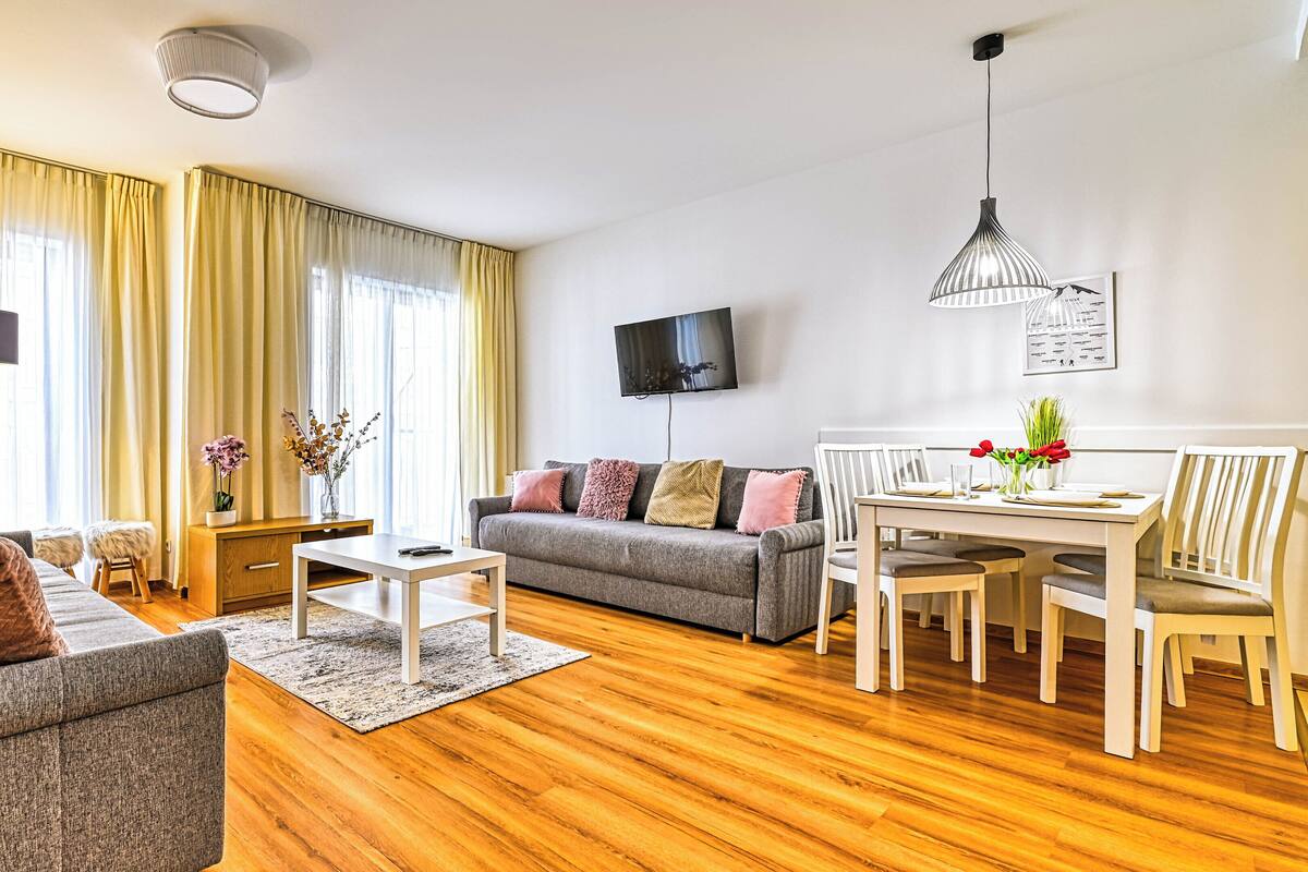 Una sala de estar moderna con un sofá, una mesa de comedor y sillas, un televisor de pantalla plana montado en la pared y una lámpara de techo.