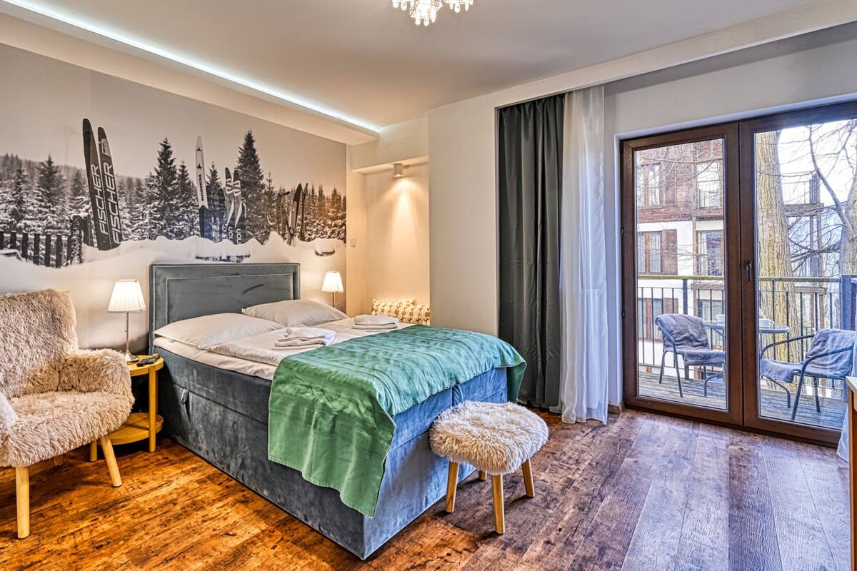 Un dormitorio con cama, una butaca mullida, una mesita de noche, una lámpara, una manta verde y un ventanal con vistas a una zona de estar exterior.