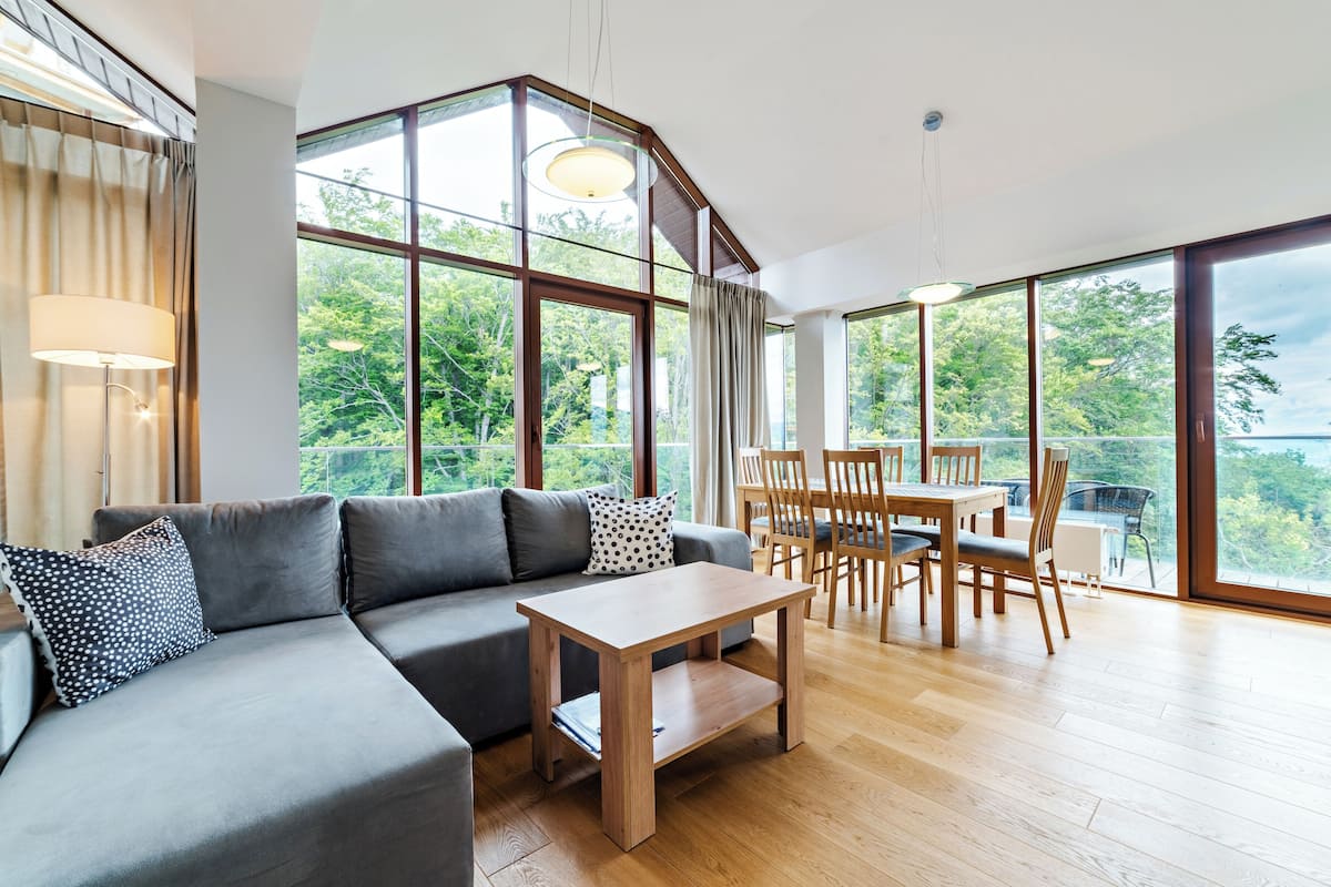Un salón moderno con un sofá gris, una mesa de centro de madera y una zona de comedor con sillas de madera. Amplias ventanas ofrecen vistas a la vegetación y un balcón.