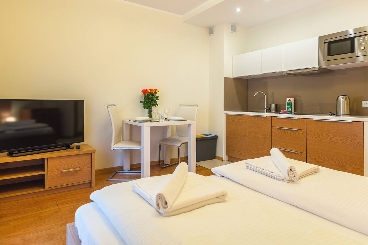 Habitación de hotel con televisión, una pequeña mesa de comedor con sillas, kitchenette y cama con toallas.