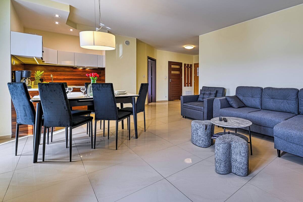 Una sala de estar moderna con un sofá gris, sillas negras, una mesita redonda y un comedor con una mesa y sillas blancas.