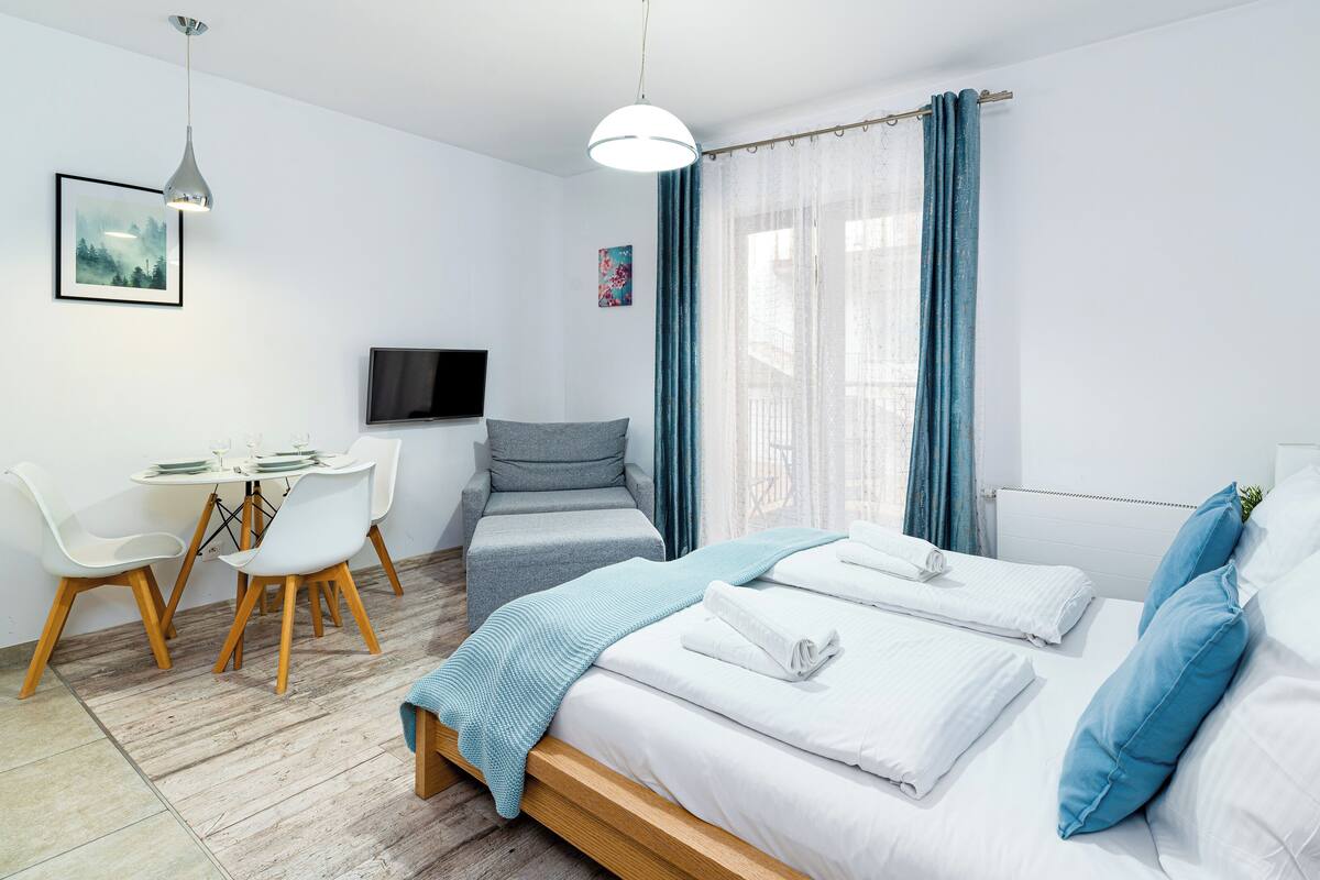 Un dormitorio moderno con cama, mesitas de noche, un escritorio con sillas, televisor y ventana con cortinas.