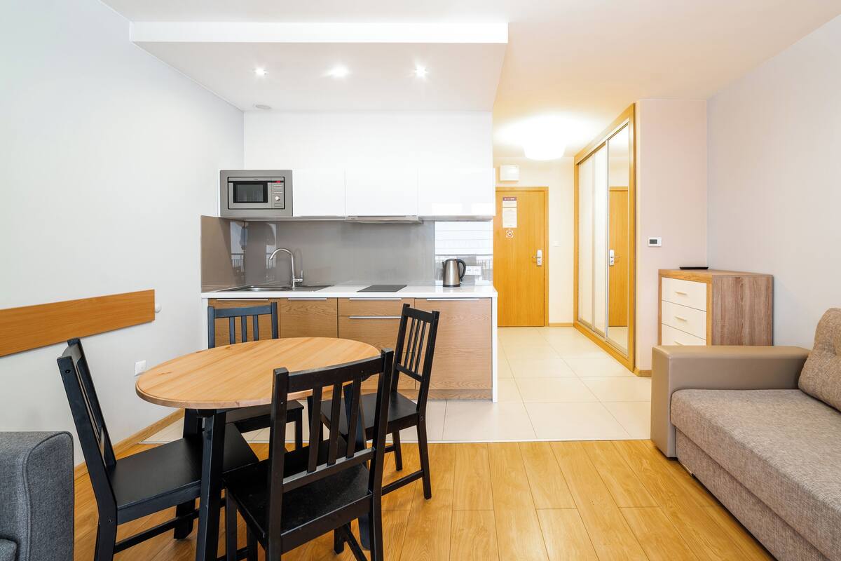 Una cocina moderna con microondas, pileta y tetera, un comedor con mesa y sillas de madera, y una sala de estar con sofá y televisor.