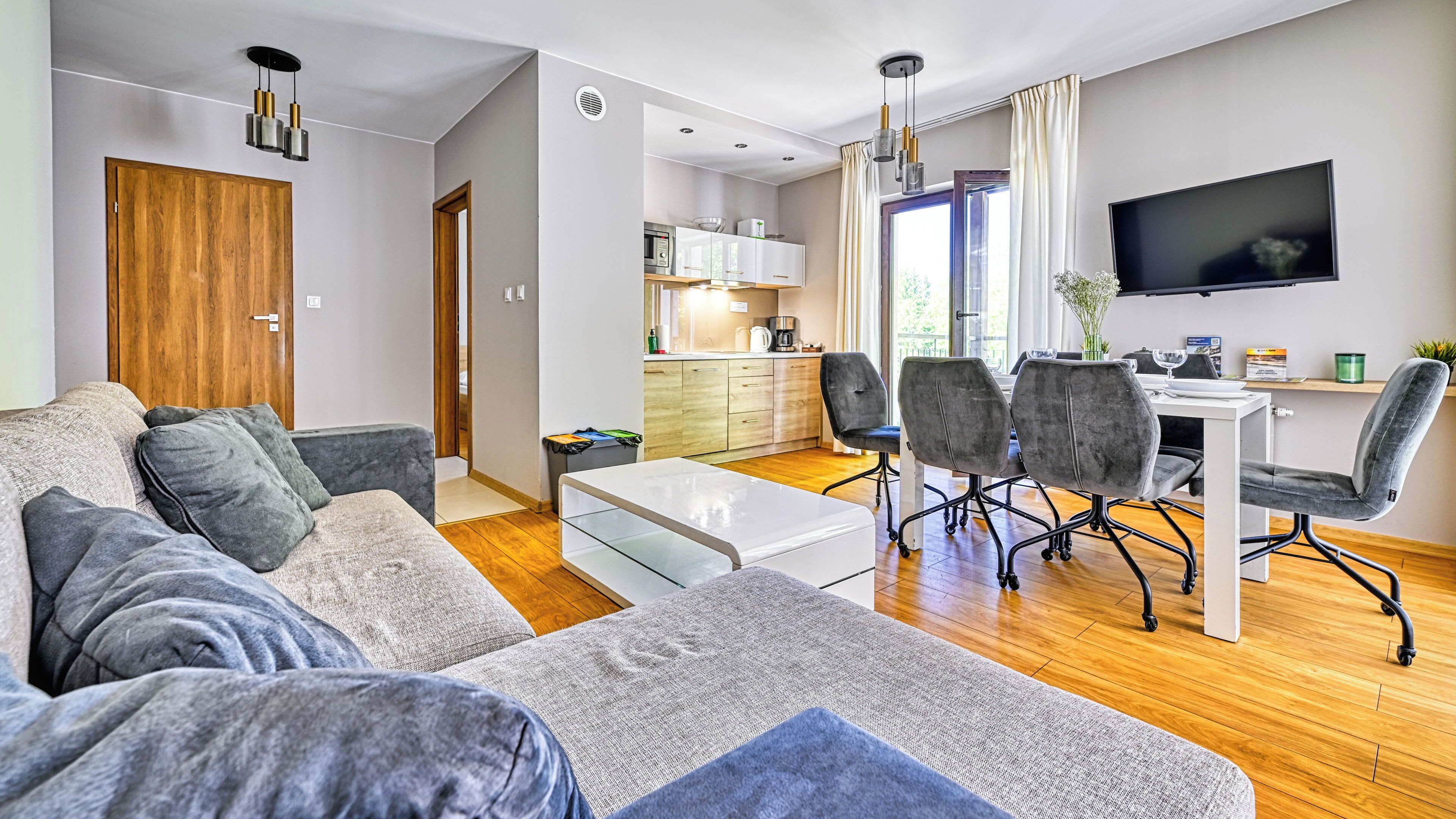 A modern living room with a sofa, a dining table with chairs, a kitchen area, and a TV mounted on the wall.