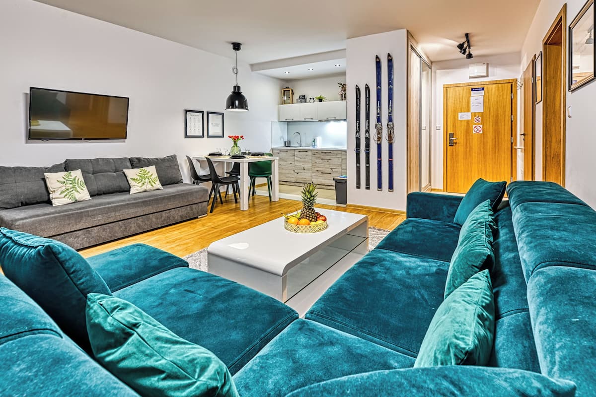 Una sala moderna con un sofá gris, sofás turquesa, una mesa de centro blanca con fruta, una cocina al fondo y una puerta de madera con equipo de esquí.