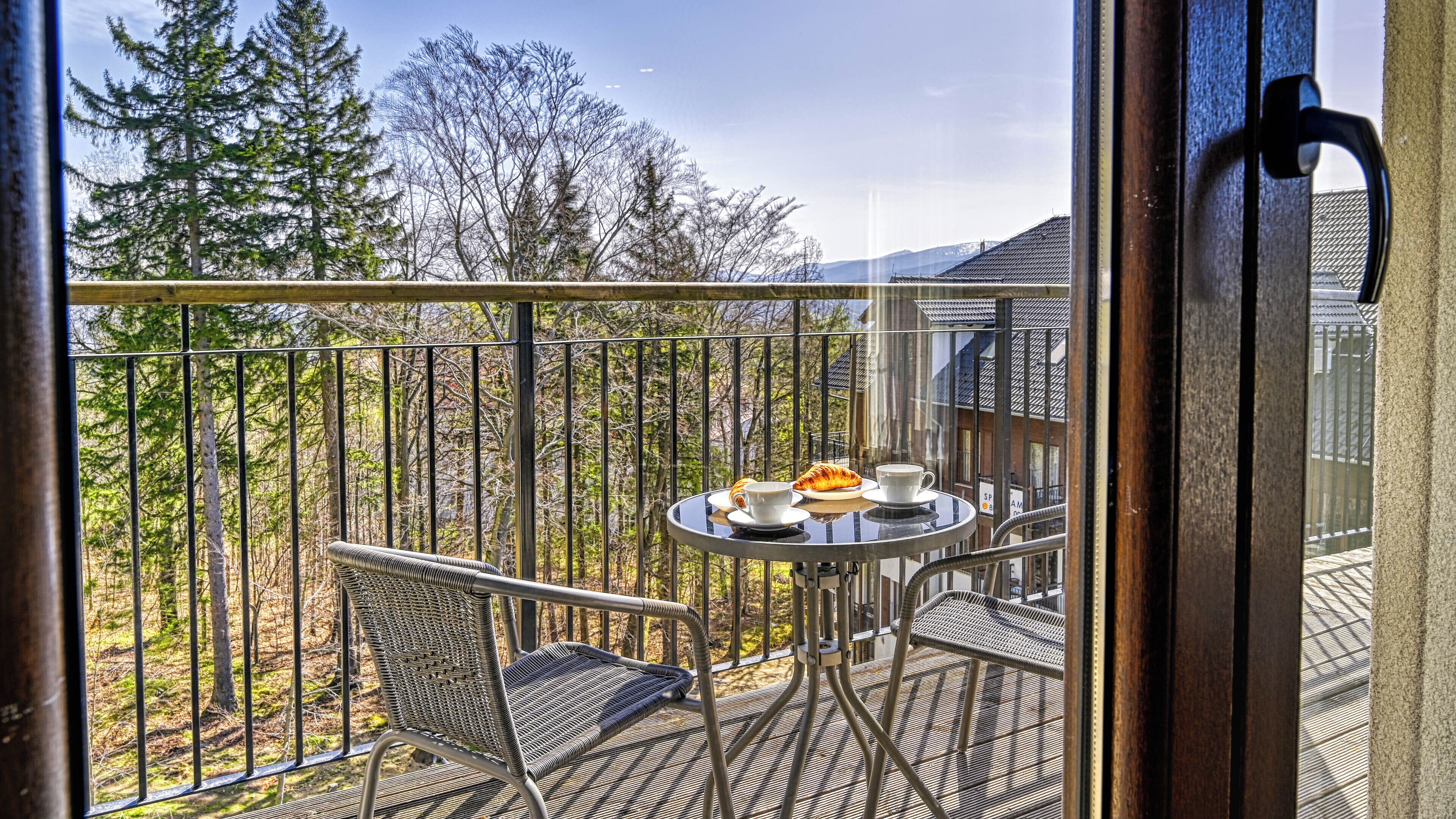 A balcony with a table set for two, featuring a croissant and a cup, overlooking a forested area.