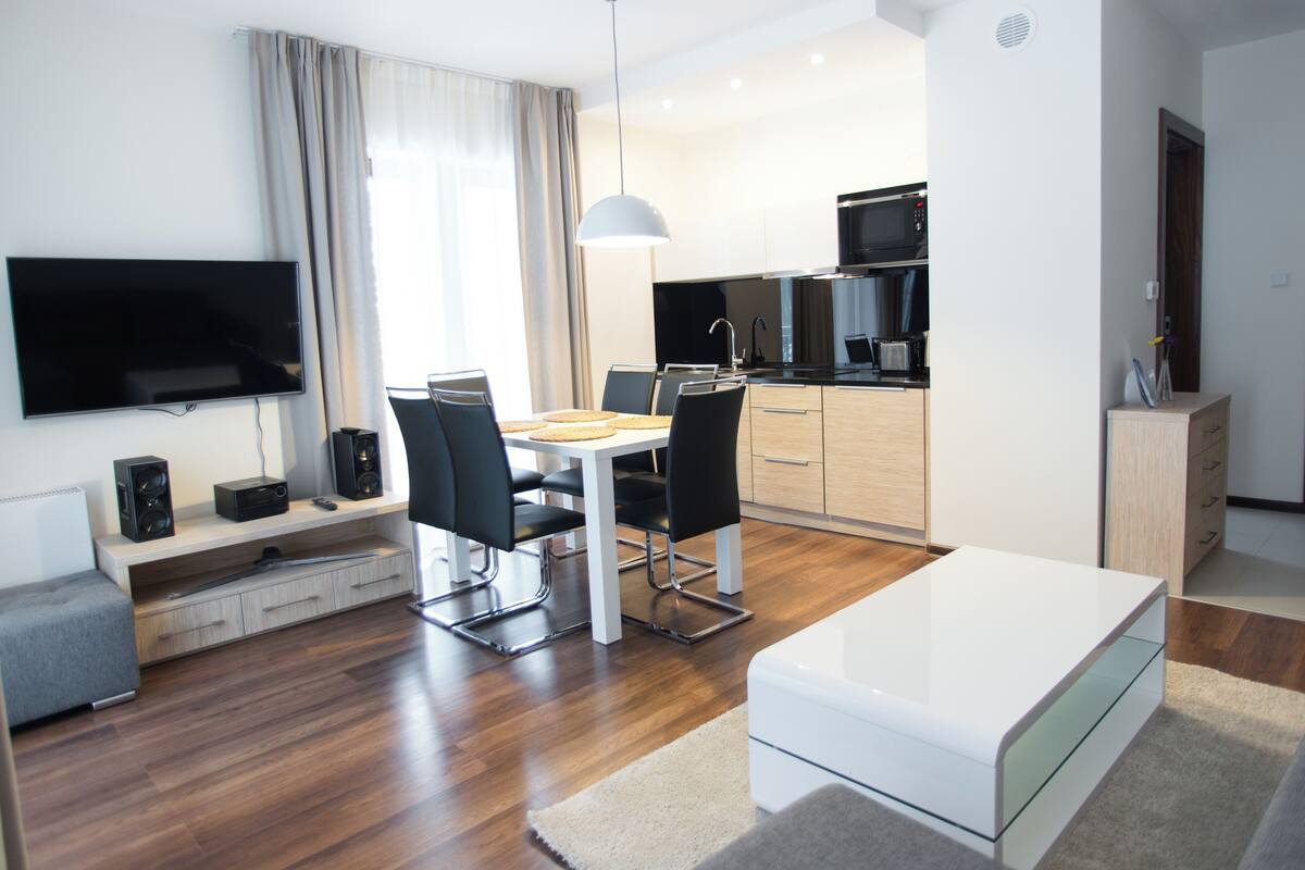 Una sala de estar moderna con televisor de pantalla plana, comedor con mesa y sillas, cocina equipada y sofá blanco.