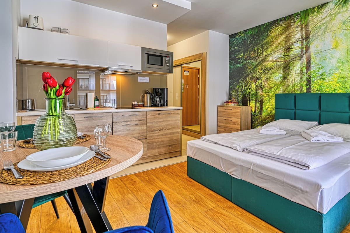 Una habitación de hotel moderna con una cama, una mesa de comedor con un jarrón de tulipanes rojos, una zona de cocina y un mural temático de bosque.