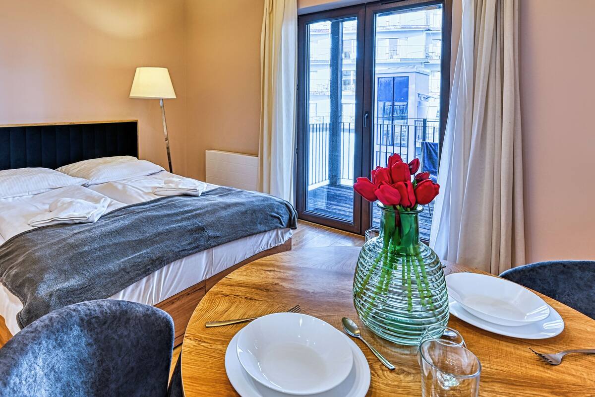 Habitación de hotel con una cama, una mesa puesta con platos, un jarrón de flores rojas y vistas a edificios a través de la ventana.