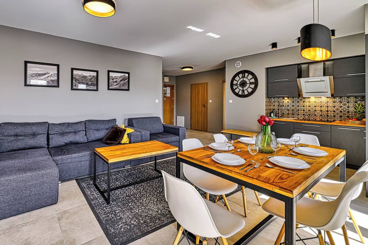 Una sala de estar moderna con un sofá gris, una mesa de comedor de madera preparada para cuatro comensales y una cocina con salpicadero de azulejos.