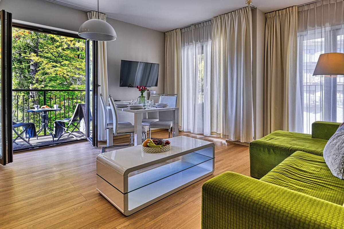 Un salón moderno con una mesa de centro de vidrio, un sofá verde, un televisor de pantalla plana y un balcón con mobiliario de exterior.