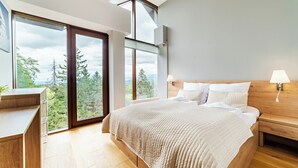 Un dormitorio con una cama grande, mesitas de noche de madera, un escritorio y vistas a árboles a través de amplios ventanales.