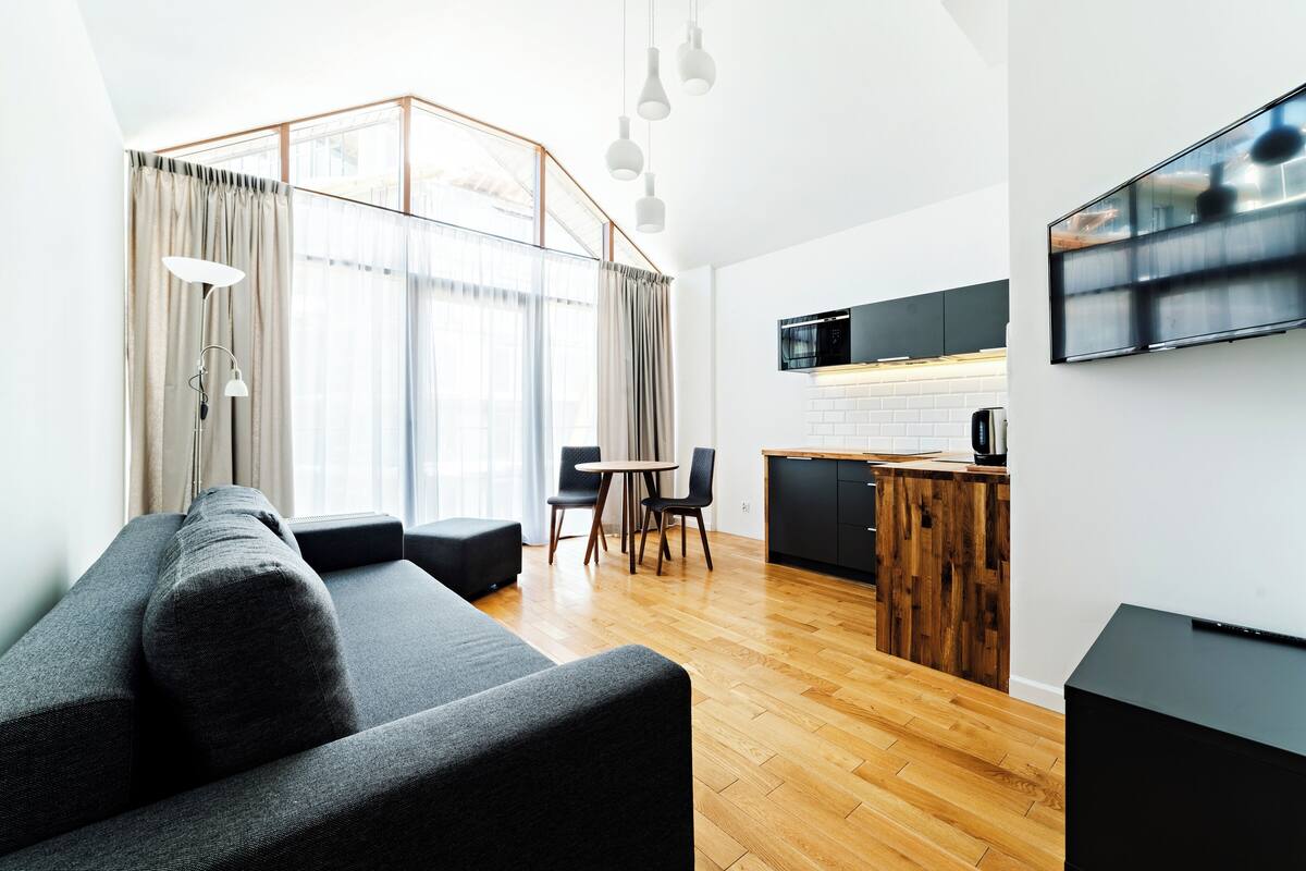 Una sala de estar moderna con sofá, mesa de comedor y sillas, un área de cocina con microondas, y un televisor montado en la pared.