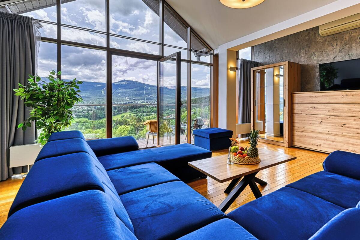 Una sala de estar moderna con ventanas amplias, un sofá azul, un mueble de madera y vistas a las montañas.