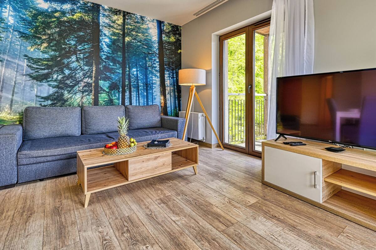 Una sala de estar moderna con sofá, mesa de centro, televisor y un mural de un bosque.