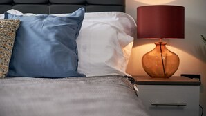 En pænt redt seng med et gråt sengegavl, en hvid pude, en blå pude og en pudse med mønster. Der er et natbord med en rød lampe og en vase.