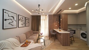 A modern living room with a sofa, coffee table, and artwork. A kitchen area with a dining table and bar stools is visible. - Designer Apartment Steps Away from Main Attractions (Yerevan)